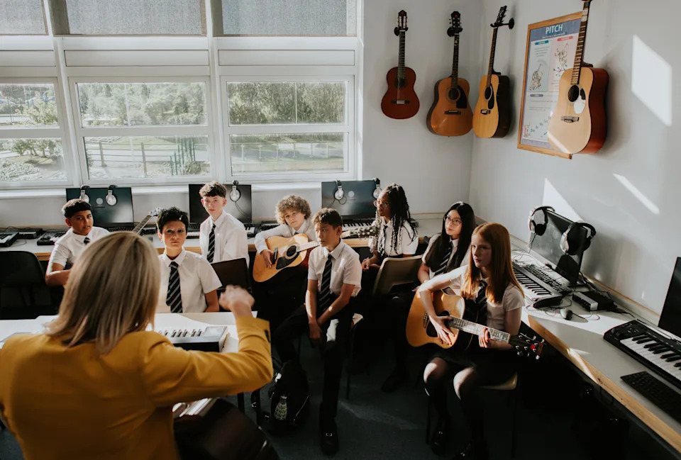 Students in uniform with guitars attend a music class, led by an instructor. Various instruments and computers are visible in the classroom