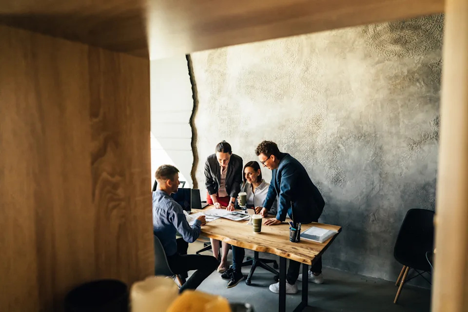 People in a meeting gather around a table discussing documents, in a modern industrial-style office