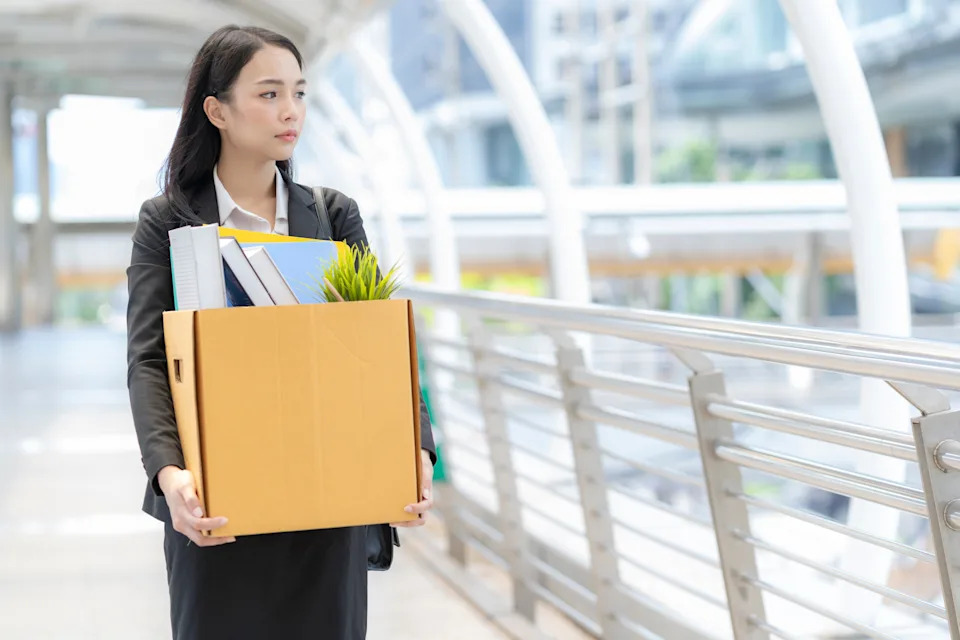 Person in business attire holding a box of office supplies, walking outdoors, possibly leaving a job