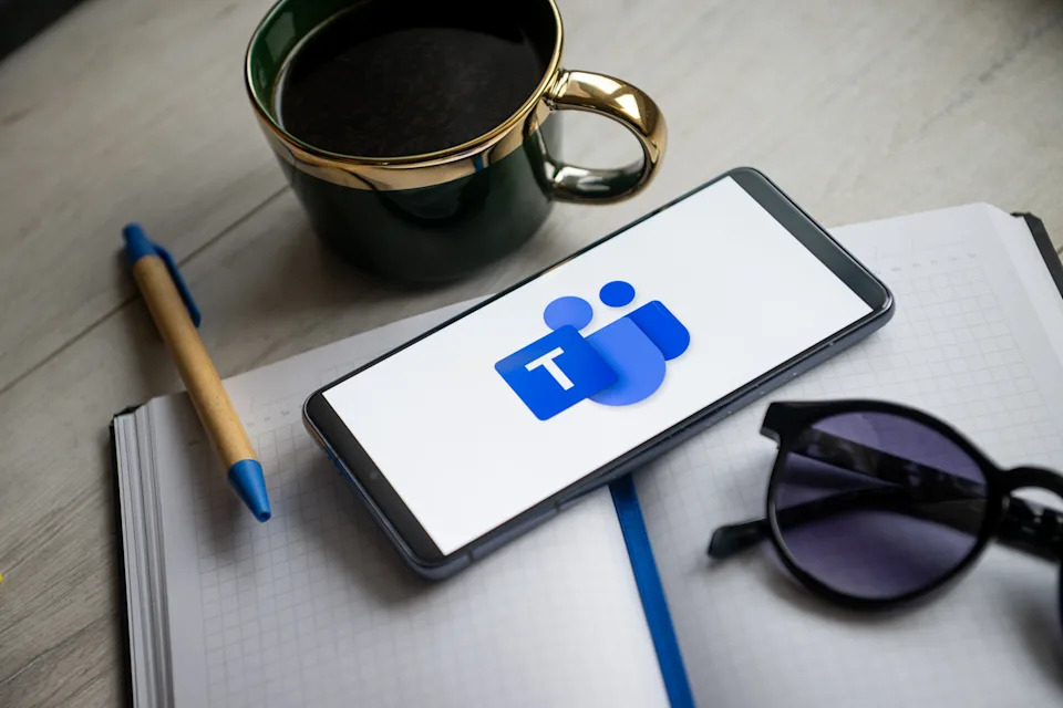 Smartphone displaying Microsoft Teams logo on notebook, beside sunglasses, pen, and coffee cup on desk