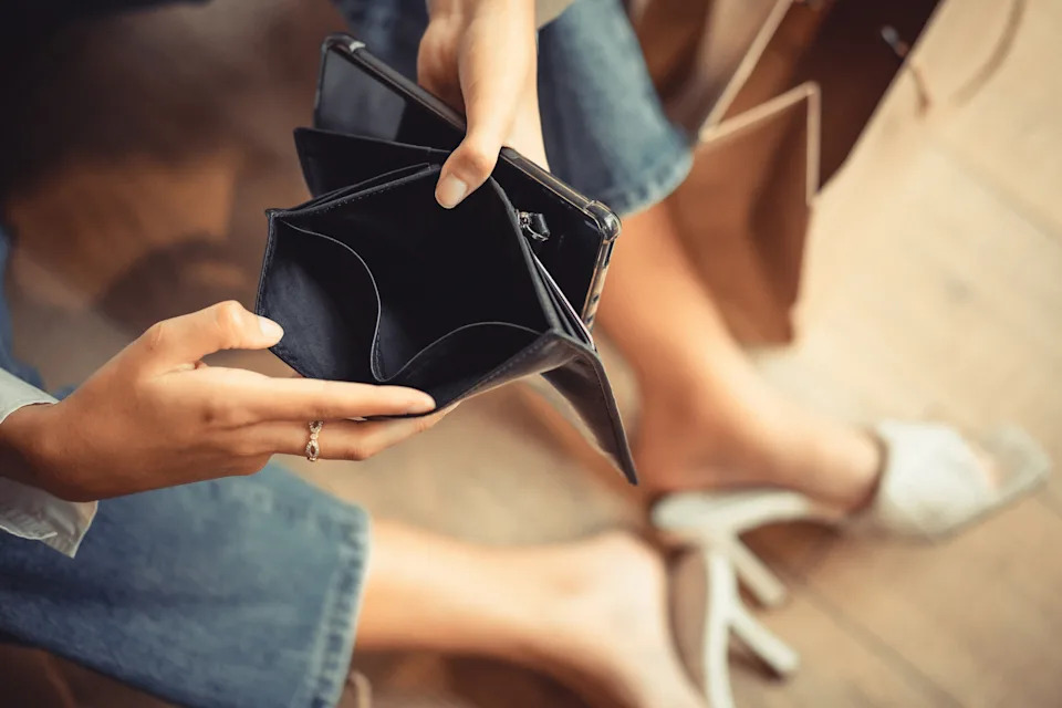 Person sitting with an empty wallet in hand, suggesting a financial topic. Casual clothing and relaxed setting indicate personal finance management