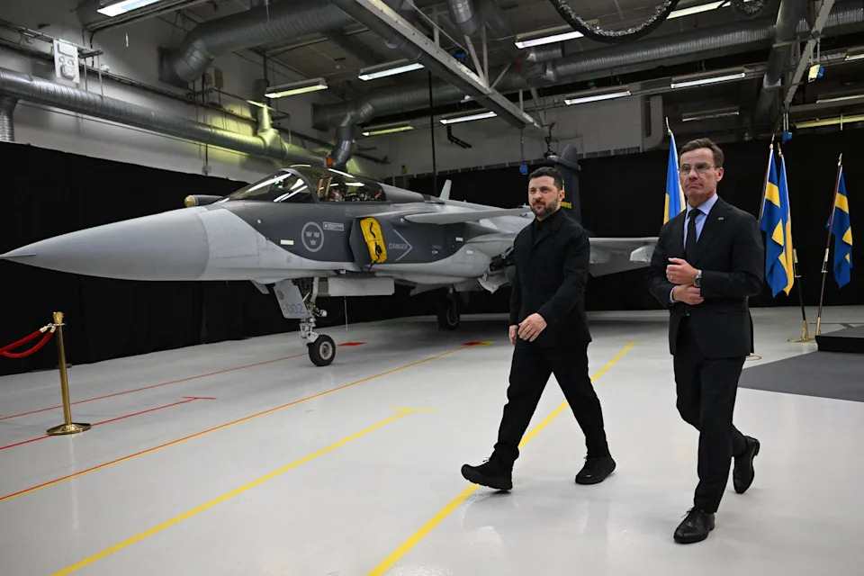 Two men walk beside a grey fighter jet inside a room
