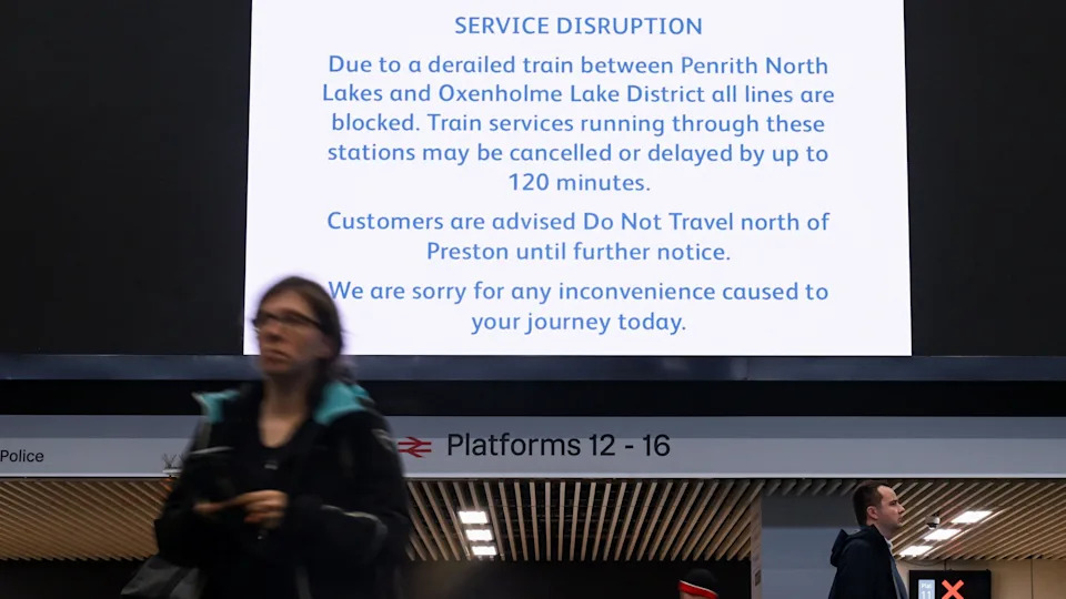 A sign at London Euston detailing the delay. There is a woman and a man walking underneath the sign. It reads 'SERVICE DISRUPTION- Due to a derailed train between Penrith North Lakes and Oxenholme Lake District all lines are blocked. Train services running through these stations may be cancelled or delayed by up to 120 minutes. Customers are advised Do Not Travel north of Preston until further notice. We are sorry for an inconvenience caused today.'